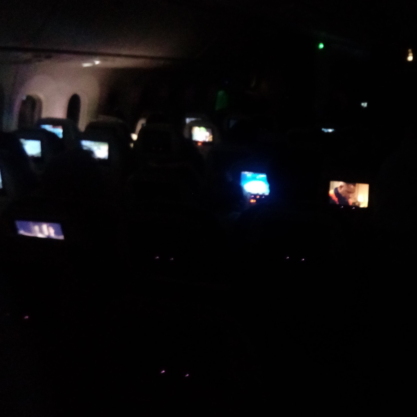 Image of interior of airliner cabin in darkness - except for numerous entertainment screens showing that people are watching movies, etc.