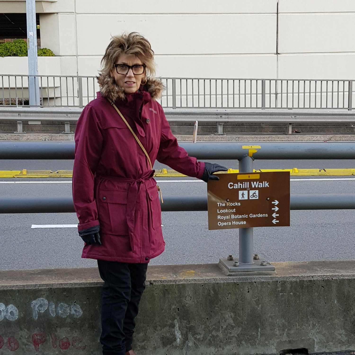 Image of Marlene wearing a warm coat, standing beside a sign on the Cahill Walkway